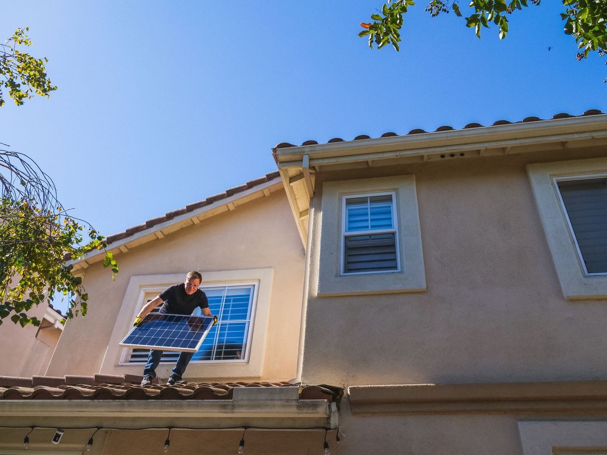 Technician on a tiled roof holding a solar panel under a clear blue sky